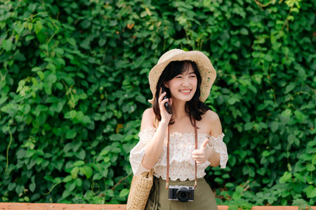 Portrait of young asian woman traveler with weaving hat, basket, mobile phone and camera on green public park background. Journey trip lifestyle, world travel explorer or Asia summer tourism concept.の写真素材