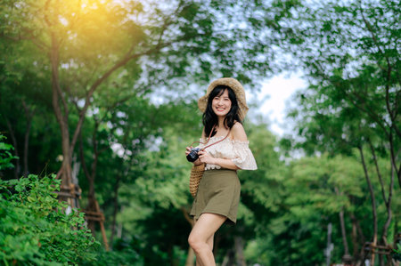 Portrait of young asian woman traveler with weaving hat and basket and a camera on green public park nature background. Journey trip lifestyle, world travel explorer or Asia summer tourism concept.の写真素材
