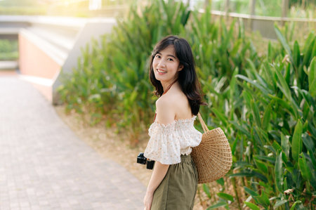 Portrait of young asian woman traveler with weaving hat and basket and a camera on green public park nature background. Journey trip lifestyle, world travel explorer or Asia summer tourism concept.の写真素材