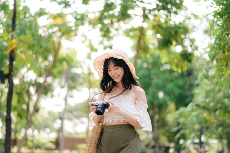 Portrait of young asian woman traveler with weaving hat and basket and a camera on green public park nature background. Journey trip lifestyle, world travel explorer or Asia summer tourism concept.の写真素材