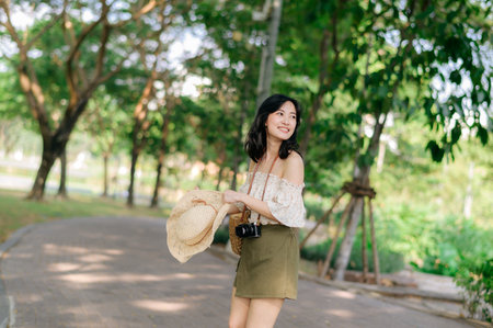 Portrait of young asian woman traveler with weaving hat and basket and a camera on green public park nature background. Journey trip lifestyle, world travel explorer or Asia summer tourism concept.の写真素材