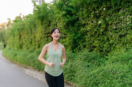 Fit young Asian woman jogging in park smiling happy running and enjoying a healthy outdoor lifestyle. Female jogger. Fitness runner girl in public park. healthy lifestyle and wellness being conceptの写真素材