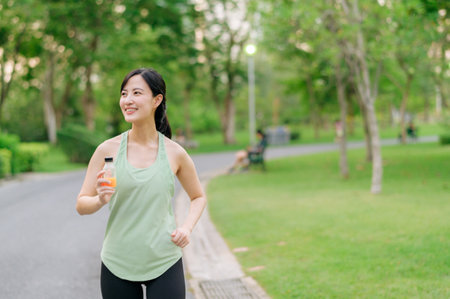 Female jogger. Fit Asian young woman with green sportswear drinking organic orange juice after running and enjoying a healthy outdoor. Fitness runner girl in public park. Wellness being conceptの写真素材