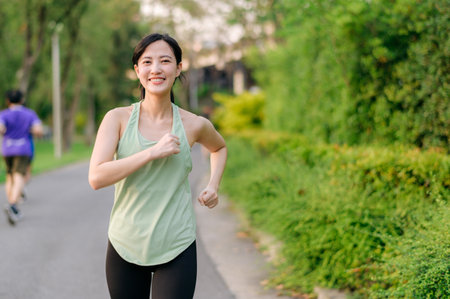 Fit young Asian woman jogging in park smiling happy running and enjoying a healthy outdoor lifestyle. Female jogger. Fitness runner girl in public park. healthy lifestyle and wellness being conceptの写真素材