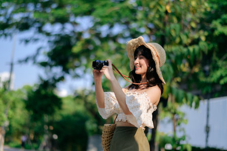 Portrait of young asian woman traveler with weaving hat and basket and a camera on green public park nature background. Journey trip lifestyle, world travel explorer or Asia summer tourism concept.の写真素材