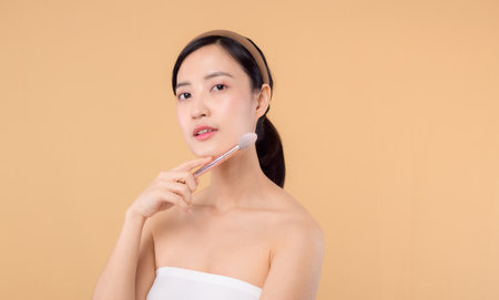 Portrait young happy asian woman with natural make up face holding cosmetic skin powder blusher isolated on beige background. Female apply skincare brush treatment. beauty product, cosmetology concept.の写真素材
