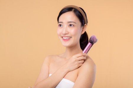 Portrait young happy asian woman with natural make up face holding cosmetic skin powder blusher isolated on beige background. Female apply skincare brush treatment. beauty product, cosmetology concept.の写真素材