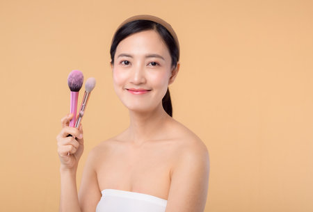 Portrait young happy asian woman with natural make up face holding cosmetic skin powder blusher isolated on beige background. Female apply skincare brush treatment. beauty product, cosmetology concept.の写真素材