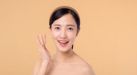 Beautiful asian girl model touching fresh glowing hydrated facial skin on beige background closeup. Beauty face young woman with natural makeup and healthy skin portrait. Skin care conceptの写真素材