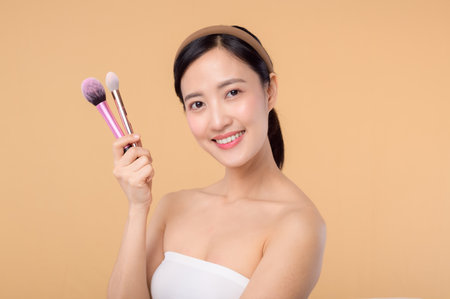 Portrait young happy asian woman with natural make up face holding cosmetic skin powder blusher isolated on beige background. Female apply skincare brush treatment. beauty product cosmetology concept.の写真素材