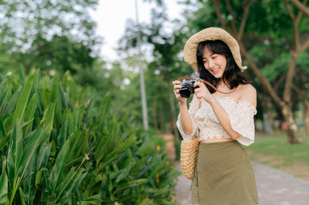 Portrait of young asian woman traveler with weaving hat and basket and a camera on green public park nature background. Journey trip lifestyle, world travel explorer or Asia summer tourism concept.の写真素材