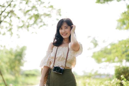 Portrait of young asian woman traveler with weaving hat and basket and a camera on green public park...