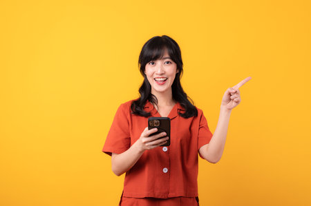 Portrait young beautiful asian woman happy smile dressed in orange clothes with smartphone and pointing finger hand gesture to free space isolated on yellow studio background. app smartphone conceptの写真素材