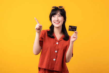 Portrait beautiful young asian woman happy smile dressed in orange clothes showing credit card and point finger to free space isolated on yellow background. Pay and purchase shopping payment concept.の写真素材
