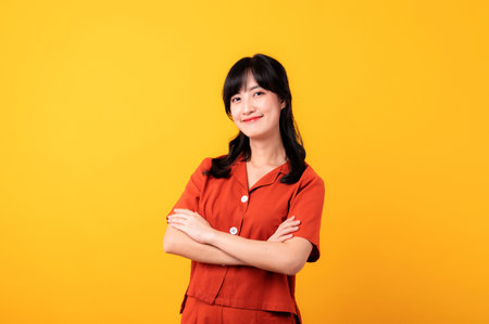 Portrait young thoughtful beautiful asian woman happy smile dressed in orange clothes showing cross arm gesture isolated on yellow studio background.の写真素材