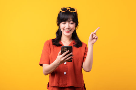 Portrait young beautiful asian woman happy smile dressed in orange clothes with smartphone and pointing finger hand gesture to free space isolated on yellow studio background. app smartphone conceptの写真素材