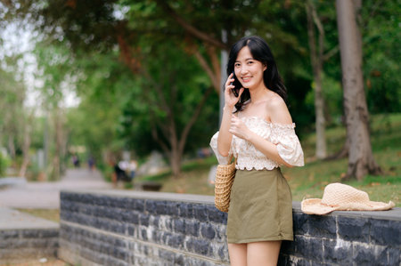 Portrait of young asian woman traveler with weaving hat, basket, mobile phone and camera on green public park background. Journey trip lifestyle, world travel explorer or Asia summer tourism concept.の写真素材