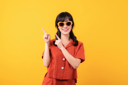 Portrait beautiful young asian woman happy smile dressed in orange clothes and sunglasses showing pointing finger hand gesture to free space isolated on yellow studio background. Attention to productの写真素材