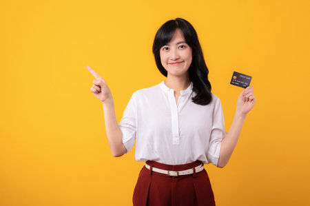 Portrait young beautiful asian woman enterpriser happy smile wearing white shirt and red plants pointing finger gesture to free copy space and holding credit card isolation on yellow background.の写真素材