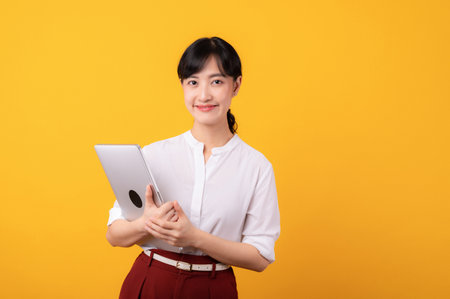 Portrait young beautiful asian woman enterpriser happy smile wearing white shirt and red plants showing confident body language gesture and holding laptop isolation on yellow background.の写真素材