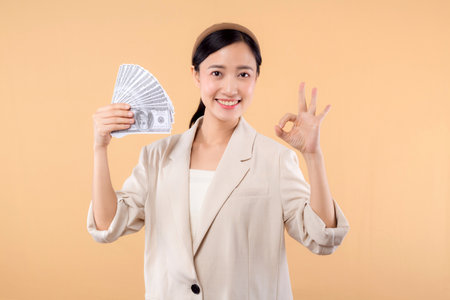 portrait of successful happy confident young asian business woman wearing white jacket holding cash money dollars standing over beige background. millionaire business, shopping concept.の写真素材