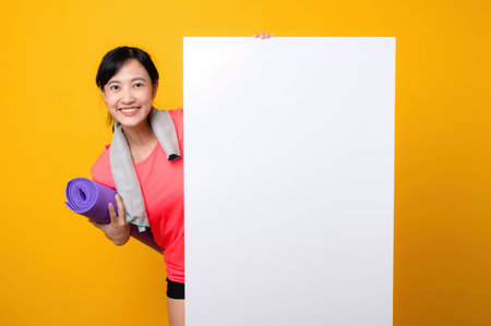 asian young sports fitness woman happy smile wearing pink sportswear standing behind the white blank banner or empty space advertisement board against yellow background. wellbeing lifestyle concept.の写真素材