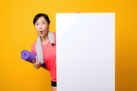 asian young sports fitness woman happy smile wearing pink sportswear standing behind the white blank banner or empty space advertisement board against yellow background. wellbeing lifestyle concept.の写真素材