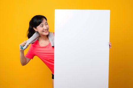 asian young sports fitness woman happy smile wearing pink sportswear standing behind the white blank banner or empty space advertisement board against yellow background. wellbeing lifestyle concept.の写真素材