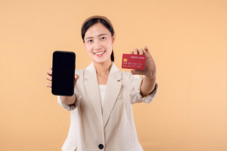 portrait of successful happy confident young asian business woman wearing white jacket holding smartphone and credit card standing over beige background. shopping concept.の写真素材