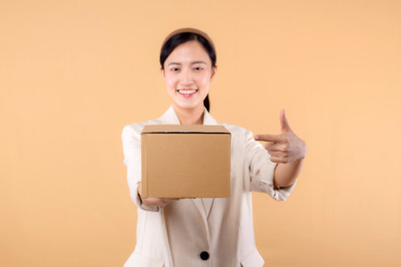 portrait of successful happy confident young asian business woman wearing white jacket holding parcel box isolated on beige studio background, Delivery courier and shipping service concept.の写真素材