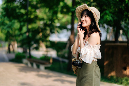Portrait of young asian woman traveler with weaving hat, basket, mobile phone and camera on green public park background. Journey trip lifestyle, world travel explorer or Asia summer tourism concept.の写真素材