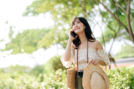 Portrait of young asian woman traveler with weaving hat, basket, mobile phone and camera on green public park background. Journey trip lifestyle, world travel explorer or Asia summer tourism concept.の写真素材