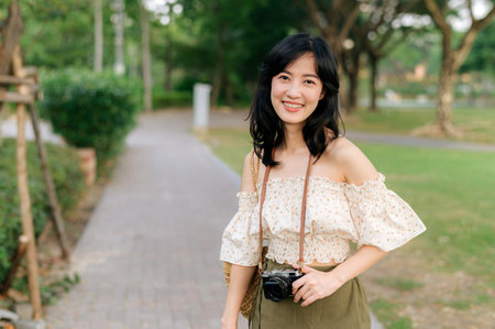 Portrait of young asian woman traveler with weaving hat and basket and a camera on green public park nature background. Journey trip lifestyle, world travel explorer or Asia summer tourism concept.の写真素材