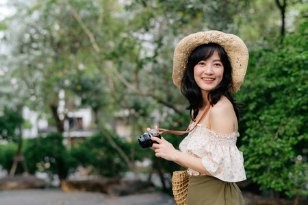 Portrait of young asian woman traveler with weaving hat and basket and a camera on green public park nature background. Journey trip lifestyle, world travel explorer or Asia summer tourism concept.の写真素材