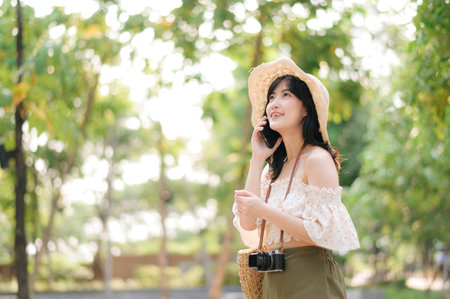 Portrait of young asian woman traveler with weaving hat, basket, mobile phone and camera on green public park background. Journey trip lifestyle, world travel explorer or Asia summer tourism concept.の写真素材