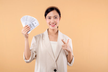 portrait of successful happy confident young asian business woman wearing white jacket holding cash money dollars standing over beige background. millionaire business, shopping concept.の写真素材