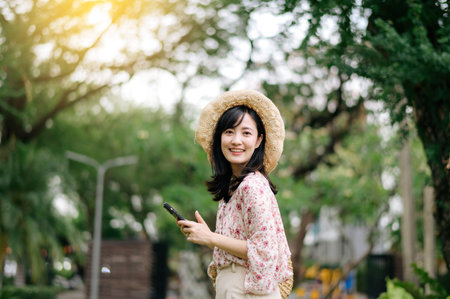Portrait of young asian woman traveler with weaving hat and basket using mobile phone on green park nature background. Journey trip lifestyle, world travel explorer or Asia summer tourism concept.の写真素材