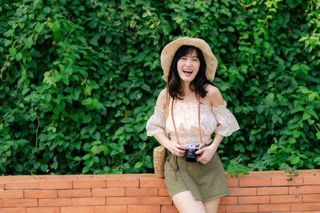 Portrait of young asian woman traveler with weaving hat and basket and a camera on green public park nature background. Journey trip lifestyle, world travel explorer or Asia summer tourism concept.の写真素材