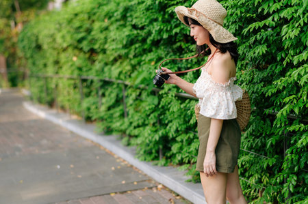 Portrait of young asian woman traveler with weaving hat and basket and a camera on green public park...