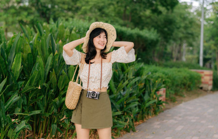 Portrait of young asian woman traveler with weaving hat and basket and a camera on green public park nature background. Journey trip lifestyle, world travel explorer or Asia summer tourism concept.の写真素材