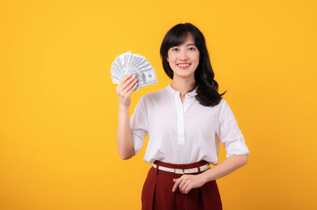 Portrait young beautiful asian woman enterpriser happy smile wearing white shirt and red plants holding cash dollar money isolation on yellow background. Money online payment concept.の写真素材