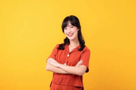 Portrait young thoughtful beautiful asian woman happy smile dressed in orange clothes showing cross arm gesture isolated on yellow studio background.の写真素材