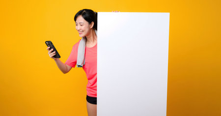 asian young sports fitness woman happy smile wearing pink sportswear standing behind the white blank banner or empty space advertisement board against yellow background. wellbeing application concept.の写真素材