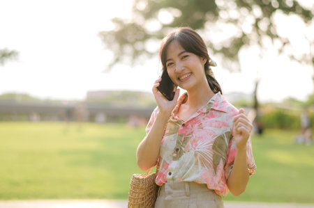 Portrait of young asian woman traveler with weaving basket, mobile phone on green public park background. Journey trip lifestyle, world travel explorer or Asia summer tourism concept.の写真素材