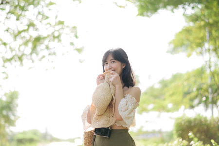 Portrait of young asian woman traveler with weaving hat and basket and a camera on green public park nature background. Journey trip lifestyle, world travel explorer or Asia summer tourism concept.の写真素材