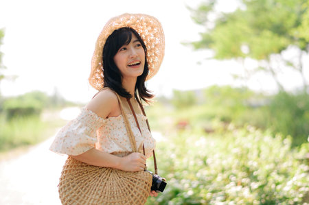 Portrait of young asian woman traveler with weaving hat and basket and a camera on green public park nature background. Journey trip lifestyle, world travel explorer or Asia summer tourism concept.の写真素材
