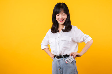 Captivating image of young Asian woman wearing white shirt and denim jeans, confidently holding cash from her shirt pocket. Fashion meets financial empowerment. Perfect for finance and fashion themes.の写真素材