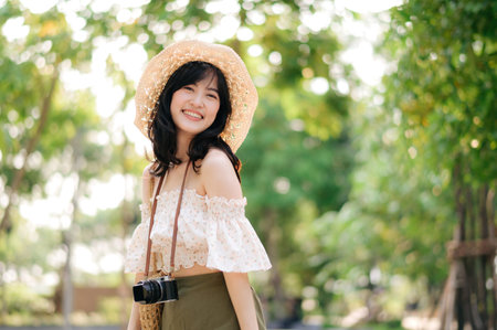 Portrait of young asian woman traveler with weaving hat and basket and a camera on green public park nature background. Journey trip lifestyle, world travel explorer or Asia summer tourism concept.の写真素材