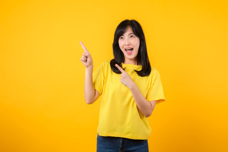 Capture attention with this stunning portrait of a confident Asian young woman wearing a yellow t-shirt and denim jeans. She points her finger to free copy space. Perfect for creative projects.の写真素材