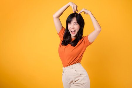 Spread love and joy with cheerful Asian woman in 30s wearing orange shirt show happy smile while forming heart shape with arms, symbolizing love and romance. Illustrating love and romantic concepts.の写真素材